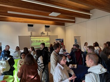 Mehrere Studierende stehen in kleinen Gesprächsgruppen zusammen. Im Hintergrund sind Stehtische, Roll-ups sowie eine Präsentation mit der Aufschrift "Herzlich willkommen" zu sehen.