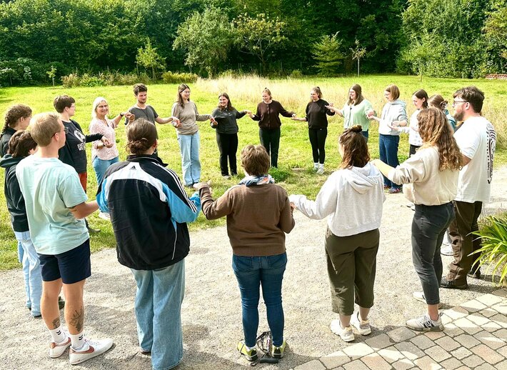 Teilnehmer_innen stehen im Kreis, schauen sich an und halten sich an den Händen.