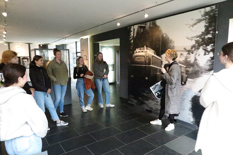 Eine kleine Gruppe Menschen steht in einem Kreis vor einem großen schwarz-weißem Bild, welches zwei alte Autos zeigt. Eine Referentin erzählt etwas über das Bild.