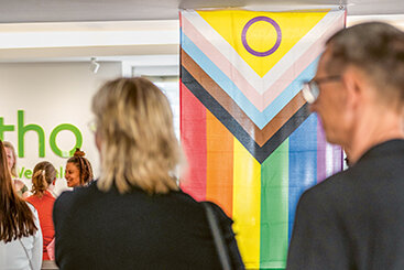 Im Foyer der katho hängt eine Progress-Flagge, eine Variation der Regenbogenflagge, die zusätzlich links einen Keil in Farben der Trans-Pride-Flagge und in braun und schwarz enthält. Im Foyer befinden sich Personen während einer Veranstaltung.