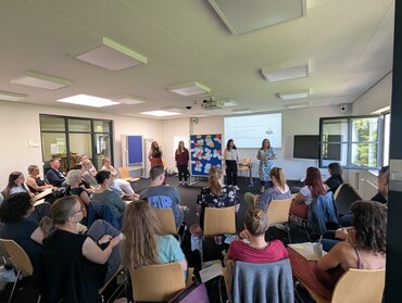 Das Bild zeigt die Mitarbeitenden im Praxisbüro, die im Raum stehen und vor den sitzenden Gästen referieren.