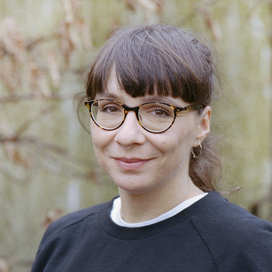 Ein Porträt Foto von Ulrike Marz. Sie hat ihre dunklen Haare zu einem Zopf zusammen gebunden. Sie trägt eine Brille und ein Septum Piercing.