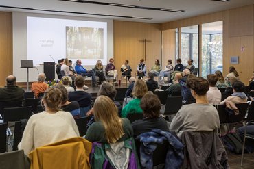 Eine große Gruppe Menschen sitzt auf dem Podium und ist im Gespräch miteinander. Im Hintergrund ist eine Präsentation zu sehen auf der steht "der Weg wohin?". Viele Menschen sitzen im Publikum und hören zu.