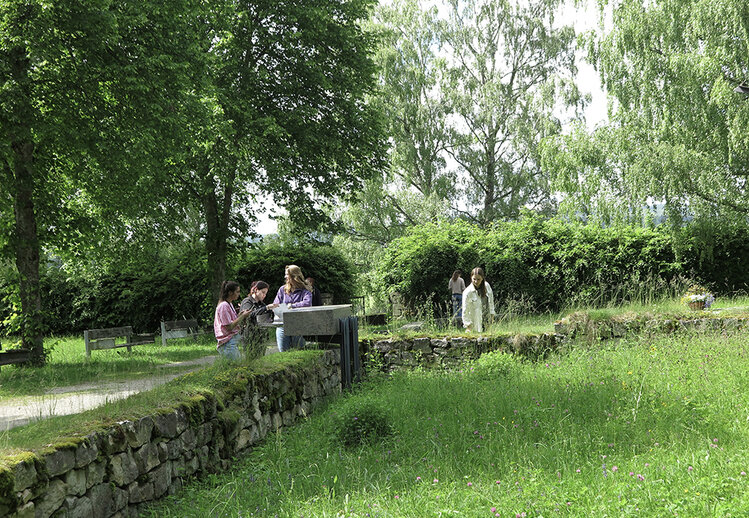 Menschen stehen in einen Garten der von einer Steinmauer umgeben ist. Teilweise blättern sie in einen Buch was in eine Steinplatte eingelassen wurde.