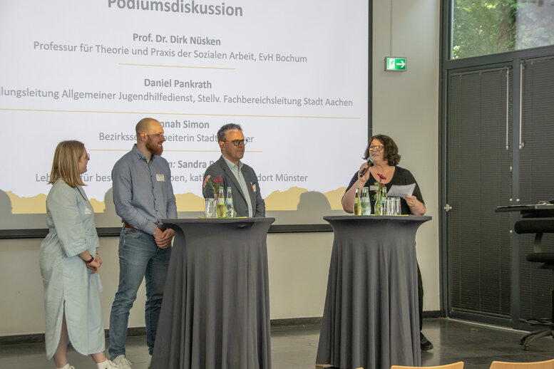 Vier Redner_innen stehen an Stehtischen auf der Bühne und diskutieren