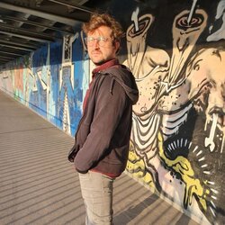 Andreas Stahl vor einer Graffitiwand. Er hat kurze blonde Haare und trägt eine Brille. Seine Hände sind in den Hosentaschen und er schaut in die Kamera.