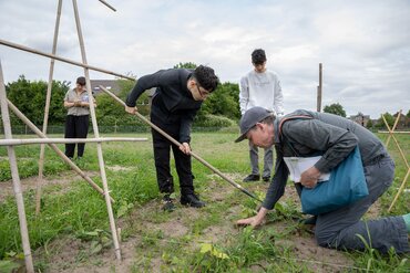 Personen erledigen Gartenarbeit, dabei benutzt einer eine Hacke. Im Hintergrund steht iene Person und scheint zu zeichnen.