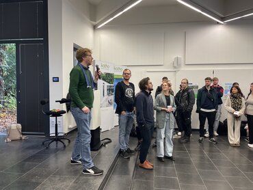 Ein Mann steht auf der Bühne mit einem Mirko in der Hand, andere Menschen hören zu.