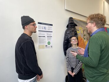 Drei Personen stehen vor einem Poster und reden miteinander.