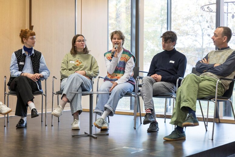 Vier Schüler*innen und ein älterer Mann sitzen auf dem Podium. Eine der Schülerinnen hält ein Mikrofon und spricht.