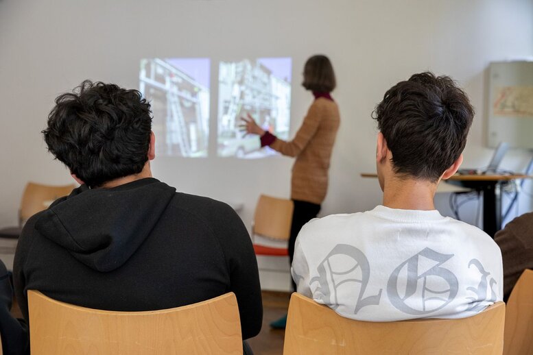 Zwei Personen schauen auf einer Leinwand, wo eine Frau einen Vortrag hält.