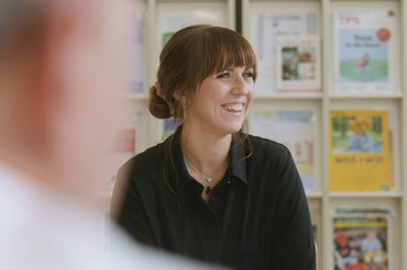 Eine Frau sitzt lachend vor einer Wand mit Zeitschriften.