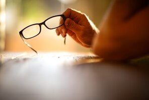 Eine Hand hält eine Brille vor einen unscharfen leuchtenden Hintergrund.