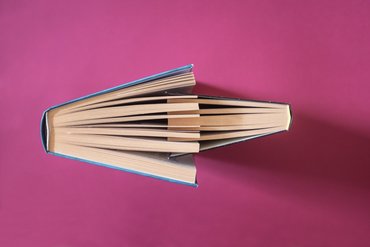 Zwei Bücher stehen sich vor einem pinken Hintergrund mit ihren Öffnungen gegenüber. Die Seiten greifen leicht ineinander über. Ein Buch ist größer. 