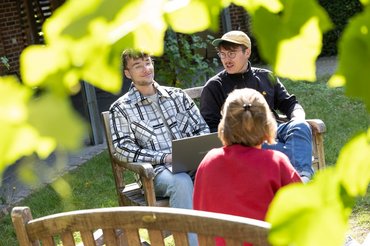 Drei Studierende sitzen zusammen auf Bänken im Grünen und unterhalten sich.