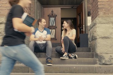 Zwei Studierende sitzen draußen auf einer Treppe und unterhalten sich. Vorne im Bild läuft eine Person vorbei.